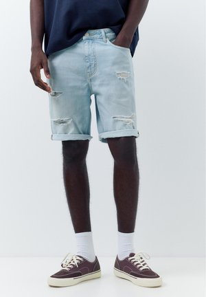 Mann trägt hellblaue, zerrissene Jeansshorts, ein dunkelblaues Hemd, weiße Socken und dunkelviolette Canvas-Sneaker vor einfarbigem Hintergrund.