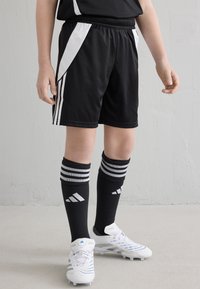 Pantalones cortos de fútbol negros con franjas laterales blancas, combinados con calcetas negras que presentan tres franjas grises, y botas de fútbol blancas con acentos azules.