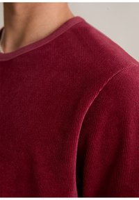 Burgundowa bluza z korduroju z okrągłym dekoltem, z prążkowaną teksturą, krótkimi rękawami i gładkim kołnierzykiem. Zbliżenie koncentruje się na okolicy ramion.