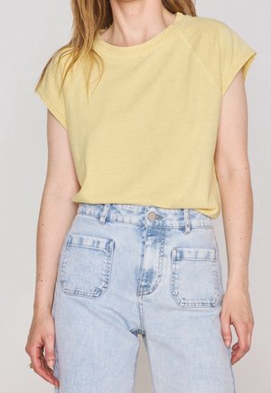 Kvinde iført en gul kortærmet top, der er stoppet ned i lyseblå denimjeans med høj talje og forlommer med lapper.
