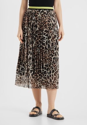 Kobieta w plisowanej midi spódnicy w panterkę z neonowo-żółto-czarnym pasem i czarnymi sandałami z klamrą, stojąca na białym tle.