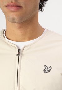 Beige zip-up jakke med jevn tekstur, med en liten brodert fuglelogo på venstre bryst. Hvit t-skjorte med rund hals under.