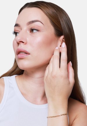 Goldene Kettenarmband am Handgelenk, Nahaufnahme der Hand in Ohrnähe. Glatte Haut mit subtiler Ausstrahlung, getragen wird ein hellgraues, geripptes Tanktop. Neutraler Nagellack.