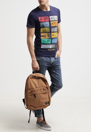 Mann trägt ein marineblaues T-Shirt mit bunten Nummernschildern, aufgerollte Jeans, graue Sneakers und hält einen braunen Rucksack seitlich.