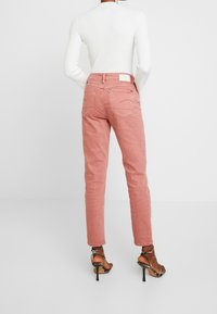 Kvinna som bär rosa raka jeans, vit långärmad topp och svarta högklackade sandaler, stående med ryggen vänd framåt.