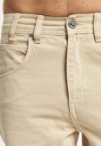 Beige cotton denim pants feature a zip fly, button closure, and standard pockets with a smooth texture and subtle diagonal weave pattern.