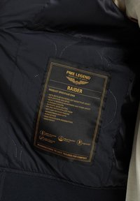 Črn prešiti notranji del lahkega pilotskega jopiča, ki vsebuje oznako za nego z zlatim tiskom, ki podrobno opisuje materiale in specifikacije izdelka.