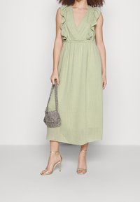 Vestido midi verde claro con escote fruncido, tejido texturizado y cinturilla elástica. Combinado con sandalias doradas y un pequeño bolso de cuentas.