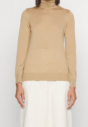 Kobieta ubrana w beżowy sweter z długim rękawem i golfem oraz białą spódnicę, stojąca na tle jednolitego białego tła, z ramionami swobodnie opuszczonymi wzdłuż ciała.