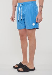 Shorts da bagno blu realizzati in materiale leggero, con cintura elastica e cordini, e un logo circolare bianco. Indossati con sandali neri.