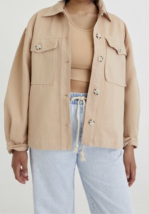 Osoba ubrana w beżową zapinaną na guziki kurtkę na dopasowany top odsłaniający brzuch i jasnoniebieskie spodnie z troczkami, stojąca na jednolitym tle.