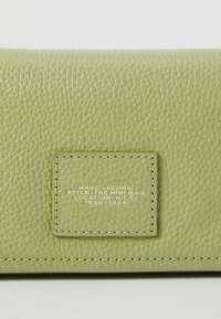 Roheline tekstureeritud nahast mini kott, millel on ristküliku kujuga plekk, kus on valge õmblusega välja toodud brändi, stiili, asukoha ja aasta andmed.