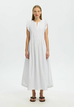 Vrouw met stijl blond haar, gekleed in een witte mouwloze midi-jurk met ruches op de schouders en zwarte sandalen, staand tegen een effen achtergrond.
