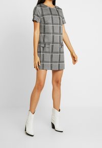 Grijs geruit jurkje met korte mouwen, twee voorzakken en een ronde halslijn, gecombineerd met witte cowboylaarzen met hak.