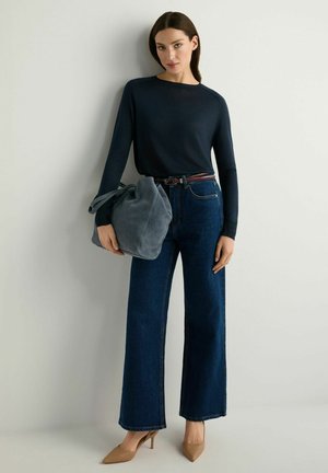 Kobieta w czarnej bluzce z długim rękawem, ciemnoniebieskich szerokich dżinsach, beżowych szpilkach, trzymająca dużą szarą torebkę, stojąca na tle gładkiej ściany.