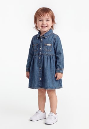 Jeune enfant souriant, portant une robe en denim bleue à manches longues et des baskets blanches, debout sur un fond blanc.