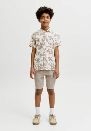 Dreng med krøllet hår iført en beige kortærmet skjorte med knapper og mønster, lysebrune shorts, hvide sokker og beige sko, stående og smilende.