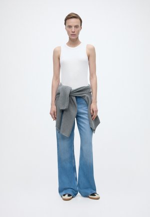 Kvinne som står iført hvit ermeløs topp, lyseblå vide jeans, grå genser knyttet rundt livet og svart-hvite joggesko.