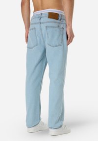 Τζιν ελαφρώς γαλάζια με χαλαρή εφαρμογή, πέντε τσέπες και δερμάτινη ετικέτα στη πίσω ζώνη μέσης. Ορατό απλό λευκό παπούτσι.