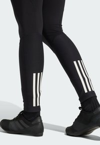 Zwarte sportleggings met drie verticale witte strepen op de onderbenen, gemaakt van rekbaar materiaal. Zwarte sportschoenen met een gestructureerd ontwerp.