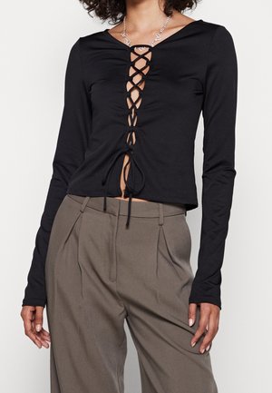 Femme portant un haut noir à manches longues avec un détail de laçage croisé sur le devant et un pantalon plissé taille haute taupe.