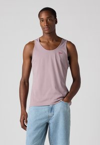 Top de alças lilás com decote redondo, apresentando um pequeno logotipo vermelho. Usado com jeans de denim azul claro, exibindo um ajuste descontraído.