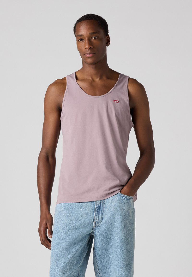 Top de alças lilás com decote redondo, apresentando um pequeno logotipo vermelho. Usado com jeans de denim azul claro, exibindo um ajuste descontraído.