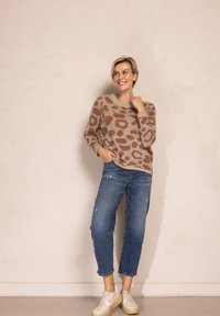 Beiger Strickpullover mit braunen abstrakten Flecken, lockere Passform, Rundhalsausschnitt, kombiniert mit hellblauen, ausgewaschenen Jeans und beigen Turnschuhen.