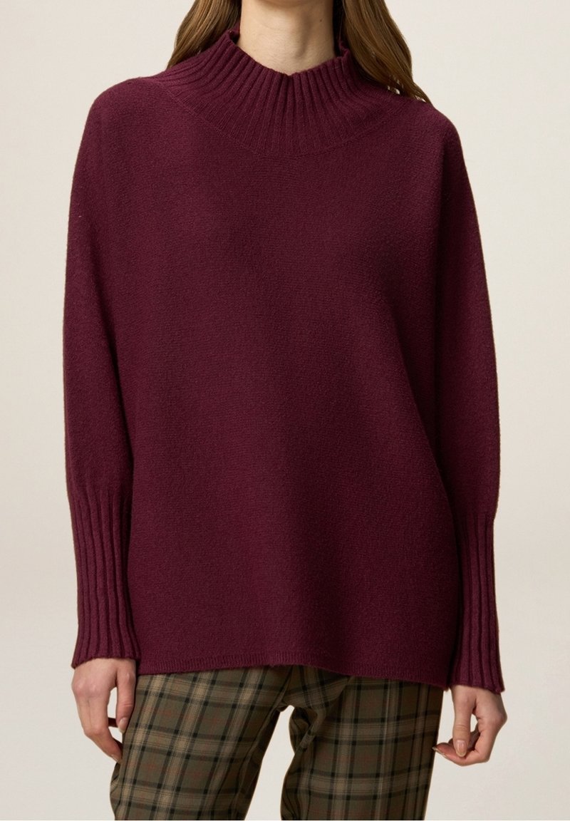 Pull en maille bordeaux avec un col montant côtelé, des épaules tombantes et des poignets côtelés texturés, associé à un pantalon à carreaux.