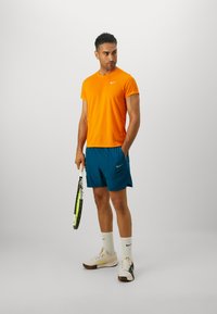 Oranžové tričko Nike, modré šortky, biele ponožky Nike a svetlé tenisky. Drží tenisovú raketu. Jednoduchý dizajn, atletický strih a kontrastné farby.