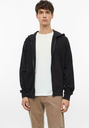 Junger Mann mit lockigem Haar, der einen schwarzen Kapuzenpullover mit Reißverschluss, ein weißes T-Shirt und hellbraune Hose vor einem schlichten weißen Hintergrund trägt.