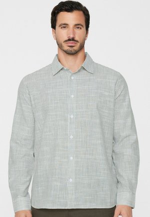 Mann trägt ein hellgrau-weiß vertikal gestreiftes langärmliges Hemd mit Knöpfen, steht vor einem einfarbigen weißen Hintergrund.