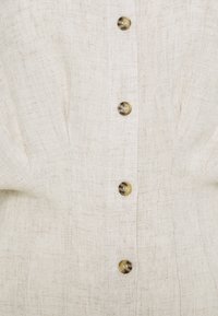 Blouse en lin de couleur beige clair avec une finition texturée, ornée de six boutons ronds foncés et de plis subtils le long de l'avant.