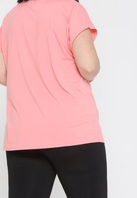 Camiseta rosa de manga corta hecha de tela ligera, con cuello redondo y un corte holgado. Se combina con pantalones negros ajustados que son visibles por debajo.