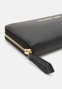 Črn teksturiran denarnica z zlatim zadrgo in dodatki, ki se ponaša z gladko površino in zapestnico za enostavno nošenje.