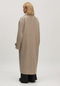 Beige Wollmantel mit lockerer Passform, seitlichen Taschen und einem einzelnen Knopfdetail, von hinten betrachtet. Glatte Textur und strukturierte Silhouette.