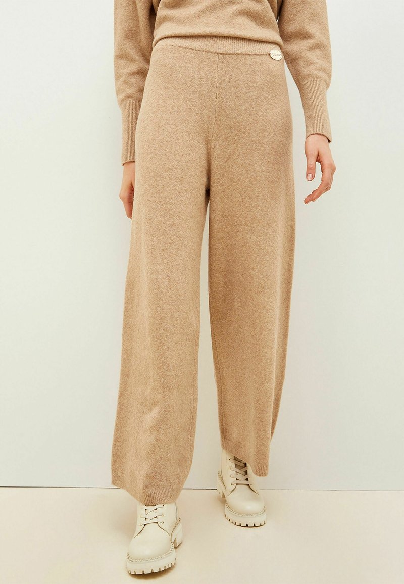 Pantalons larges beiges en tissu doux et texturé, taille haute avec un détail de bouton décoratif sur le côté. Associés à des bottes blanches.