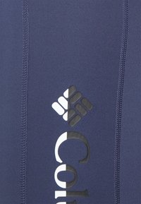 Logo Columbia vertical noir et blanc sur tissu bleu foncé avec des coutures visibles des deux côtés.