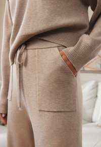 Beežist kudumiga kampsun soonikkoes äärega, millele on lisatud sobivad püksid, millel on nöörvöökoht ja esitaskud. Pehme tekstuur, lõdvestunud istuvus.