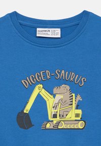 Μπλε μπλουζάκι για νήπια με ένα κινούμενο σχέδιο δεινοσαύρου που οδηγεί ένα κίτρινο εκσκαφέα και το κείμενο "DIGGER-SAURUS" πάνω από το σχέδιο.
