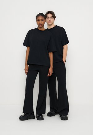 Δύο νέοι ενήλικες στέκονται, φορώντας ίδια φαρδιά μαύρα μπλουζάκια, μαύρο παντελόνι και χοντρά μαύρα παπούτσια, με φόντο ένα απλό λευκό φόντο.