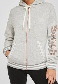 Grijze zip-up hoodie met een capuchon, witte koordjes, zijzakken en zilveren accenten. Heeft gestreepte boorden en een logo van rosegouden studs.