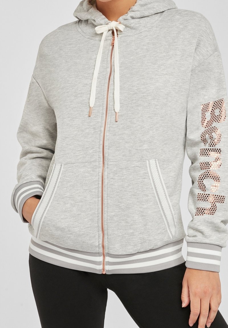 Grijze zip-up hoodie met een capuchon, witte koordjes, zijzakken en zilveren accenten. Heeft gestreepte boorden en een logo van rosegouden studs.