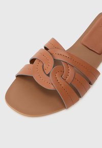 Sandalia plana marrón con diseño de correas trenzadas, textura suave y forma de punta redondeada con detalles de costura gris.