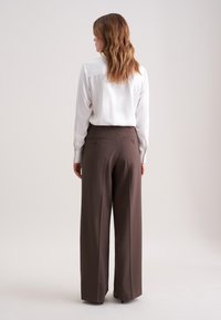 Bruine wijde broek met een gladde textuur, featuring twee achterzakken en een knoopsluiting. Gecombineerd met een witte shirt met verticale strepen.
