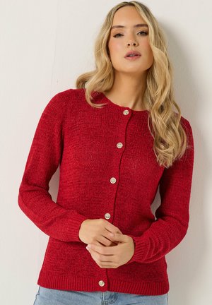 Frau mit langen blonden Haaren, die eine rote, geknöpfte Strickjacke und hellblaue Jeans trägt, steht vor einem neutralen Hintergrund.