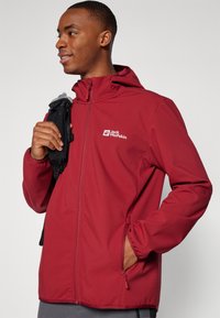 Rød softshell-jakke med hætte, lynlås foran og sidelommer. Har et logo på brystet og en glat, strækbar tekstur.
