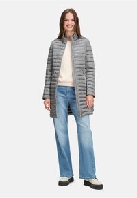 Smilende kvinde iført en lang grå quiltet jakke, cremet sweater, lyseblå jeans og hvide plateau sko, stående foran en hvid baggrund.