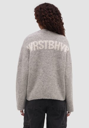 Femme aux longs cheveux bouclés portant un pull gris duveteux avec des lettres blanches sur le haut du dos et un pantalon noir.