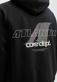 Sort hættetrøje med et stort grafik, der viser teksten "ATLANTIC" og "core dept." i hvide og lilla nuancer, lavet af et blødt bomuldsblandingsmateriale.